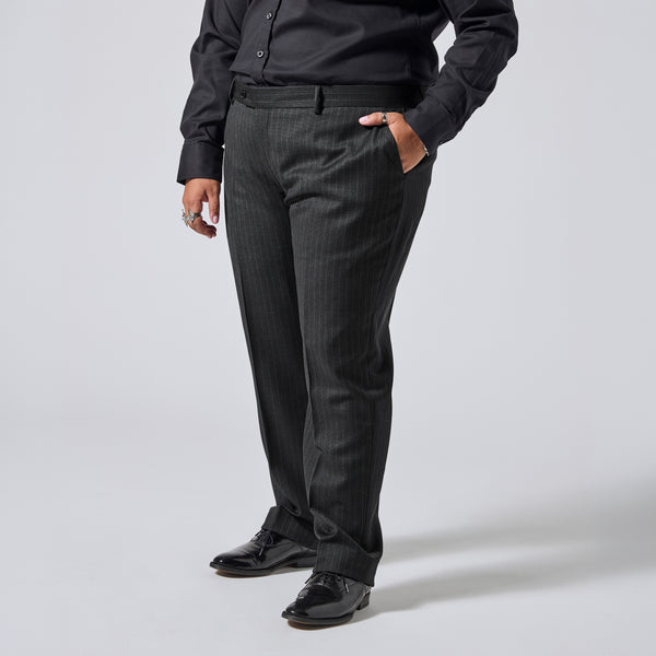 A person wearing dark charcoal gray pinstriped dress pants with a black shirt and black dress shoes, standing against a light gray studio background. Color: Shadow Pinstripe