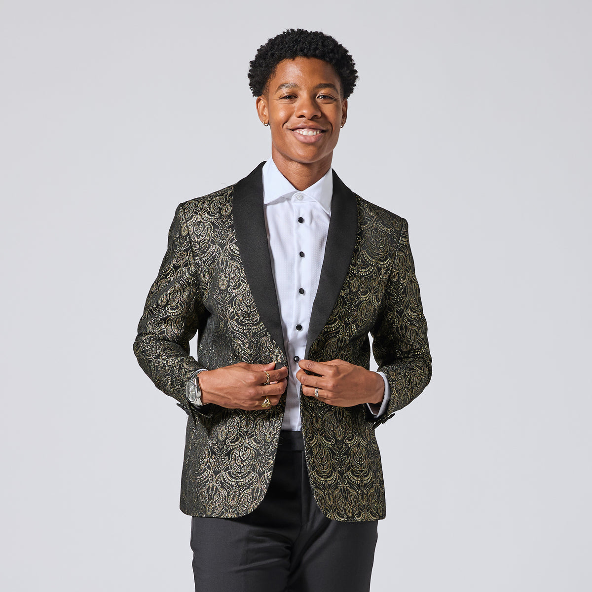 Model wearing a slim-fit blazer with a black and gold damask pattern, black satin shawl collar, and black trousers, posing against a light gray studio background. Color: Gilded Brocade
