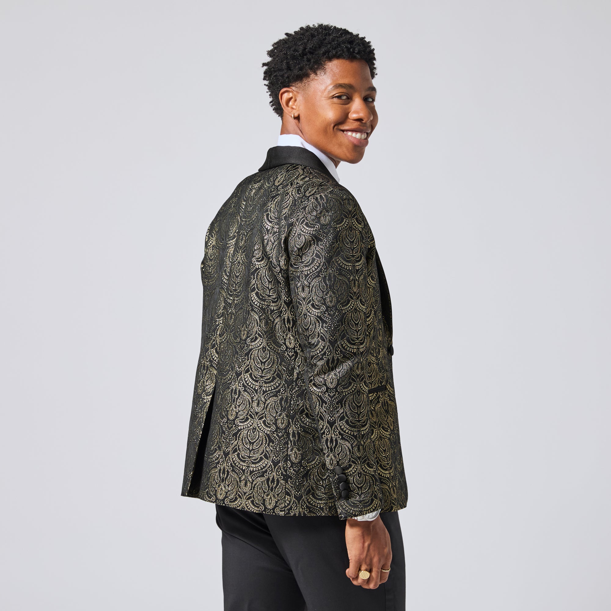 Model wearing a black and gold patterned blazer, shown from a three-quarters back view against a light gray studio background. Color: Gilded Brocade