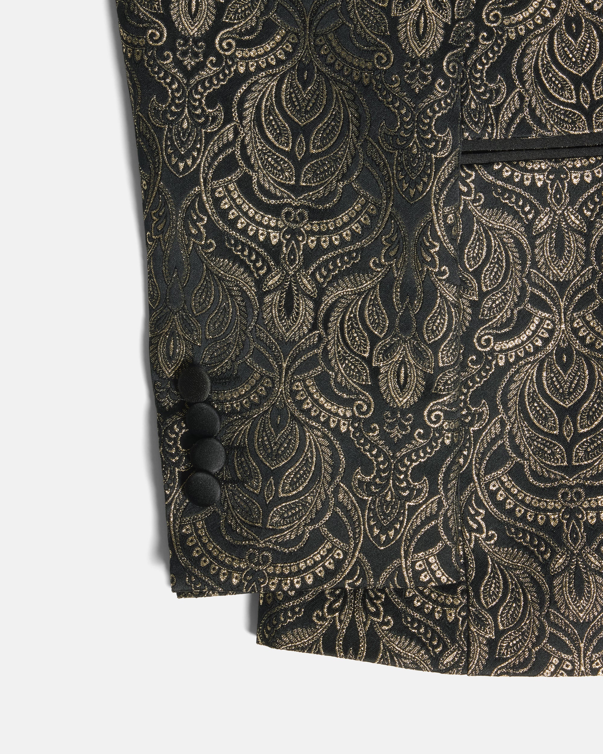 Close-up of a black and gold jacquard fabric garment, showing an ornate paisley pattern, four black buttons on the sleeve cuff, and a pocket edge against a light gray background. Color: Gilded Brocade