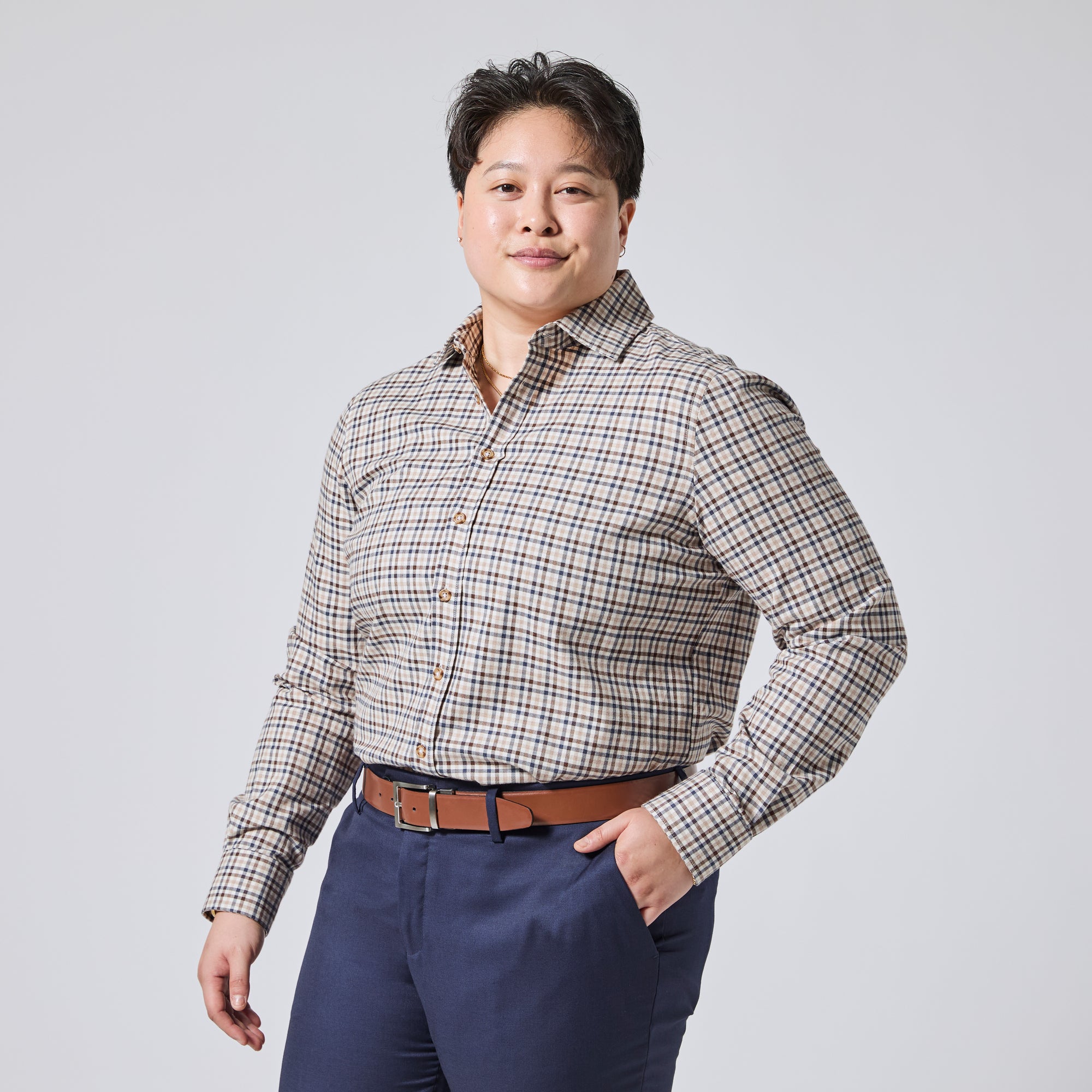 Model wearing a beige and navy blue plaid button-down shirt with a brown belt and navy trousers, standing against a light gray studio background. Color: Herritage Plaid