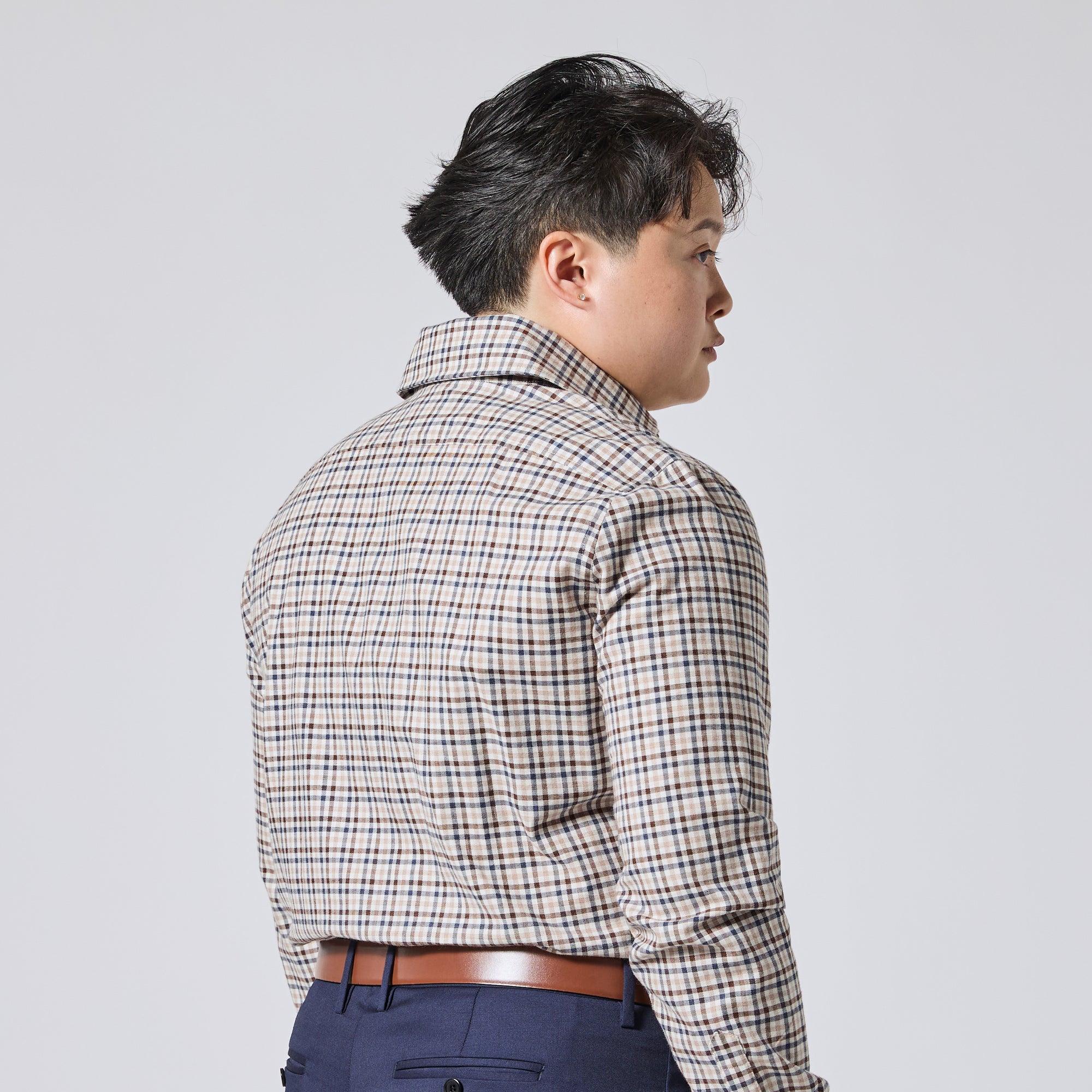 Model shown from the back and side wearing a beige plaid button-down shirt with dark blue trousers and a brown belt, photographed against a solid light gray background. Color: Herritage Plaid