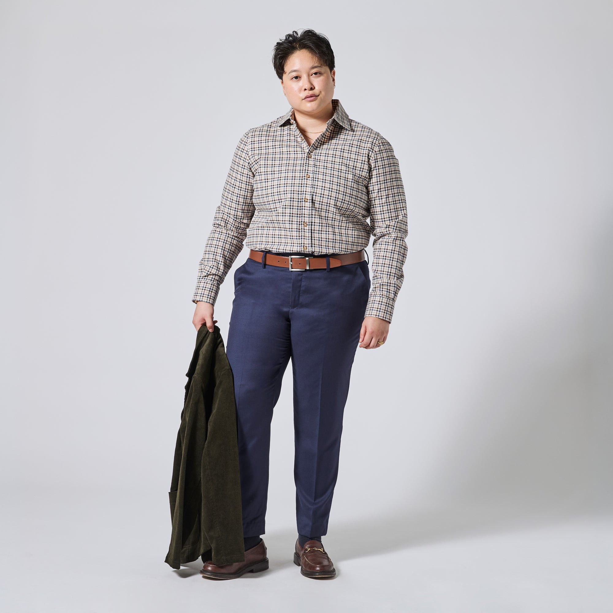 Person modeling a long-sleeve button-up plaid shirt in beige, brown, and gray, paired with navy blue trousers, a brown leather belt, and brown shoes. They are holding an olive green jacket against a light gray studio background. Color: Herritage Plaid