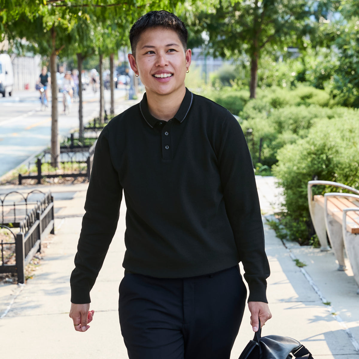 A smiling model walking outdoors on a sunny sidewalk wearing a black long-sleeve polo shirt and dark pants, holding a black bag. Color: Gray Tipped