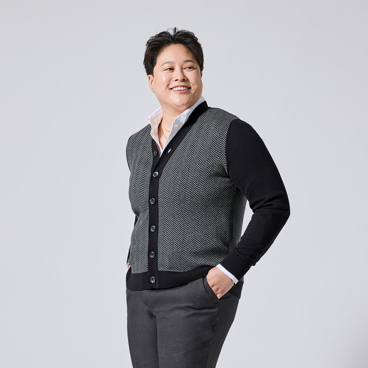 Person modeling a black and gray herringbone pattern cardigan vest over a black long-sleeve shirt and white collared undershirt, paired with gray trousers, against a light gray studio background. Color: Inkstone Herringbone