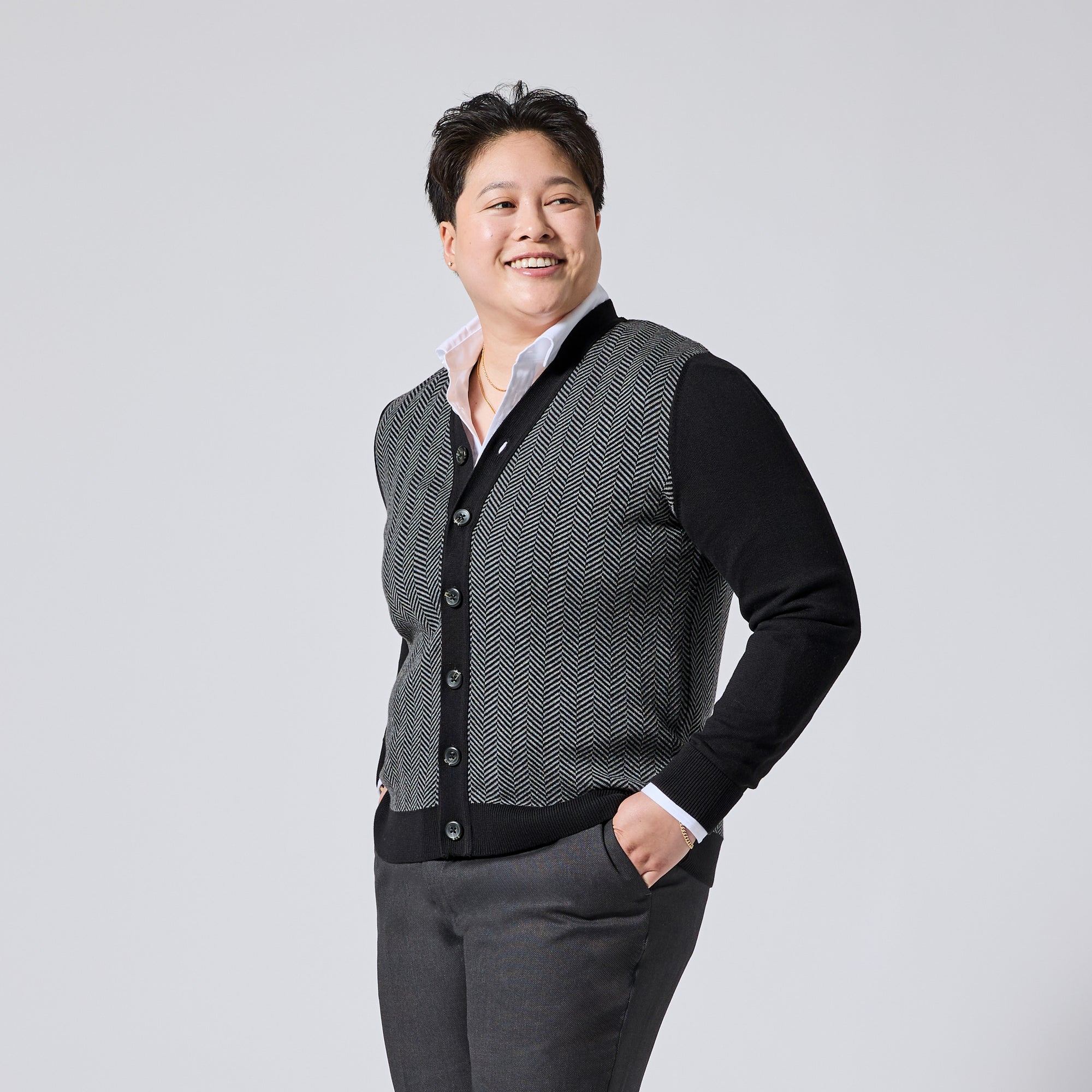 Person modeling a black and gray herringbone pattern cardigan vest over a black long-sleeve shirt and white collared undershirt, paired with gray trousers, against a light gray studio background. Color: Inkstone Herringbone