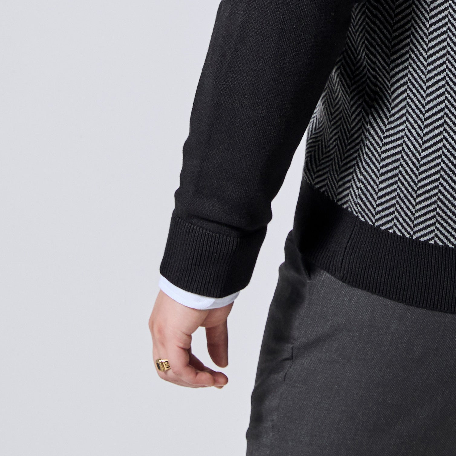 Close-up of a person wearing a black knit sweater with a black-and-white herringbone pattern on the chest, paired with dark gray trousers and a white collared shirt cuff visible at the sleeve. Color: Inkstone Herringbone