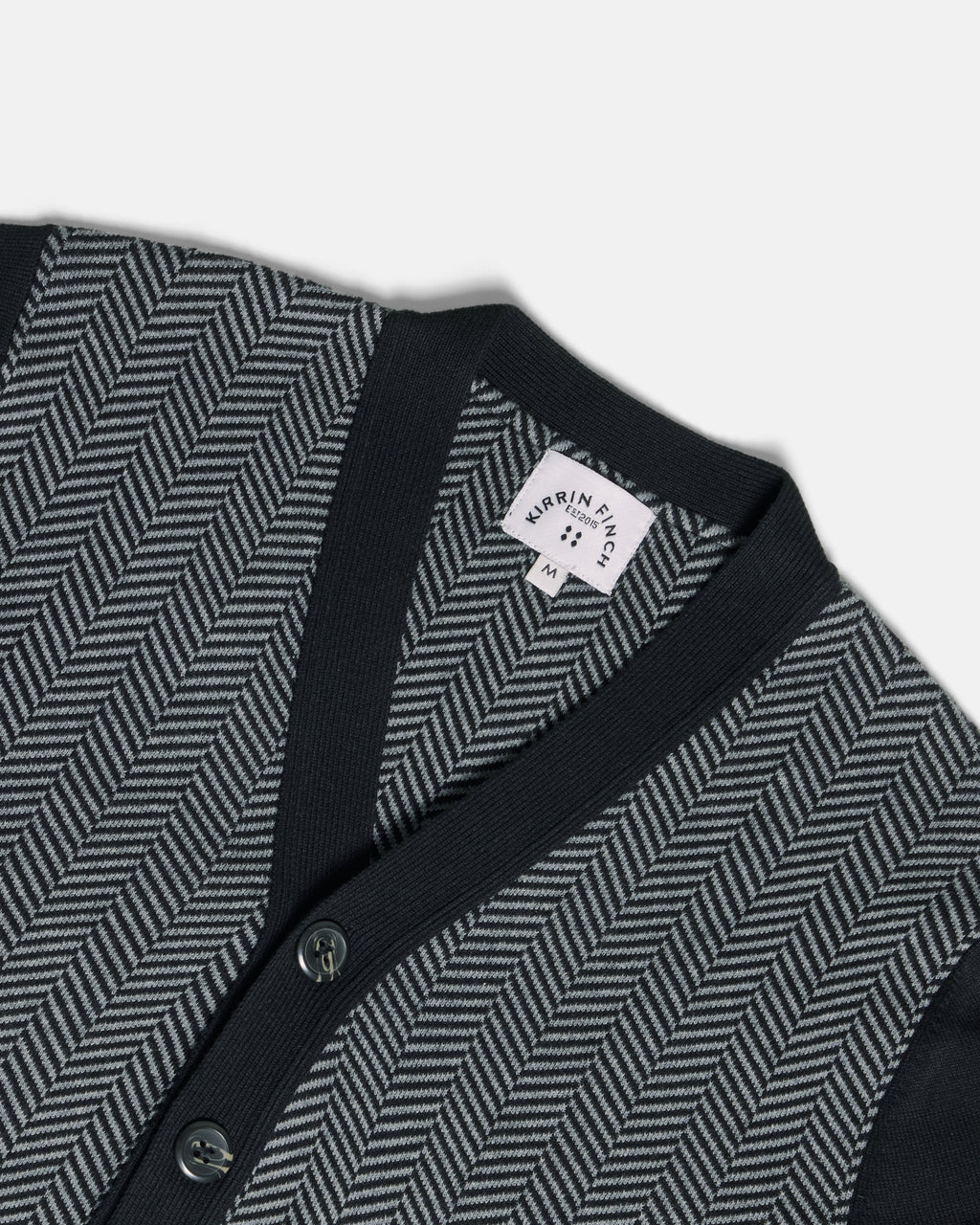 Close-up of a knit cardigan with a dark navy V-neck and button placket, paired with a gray and black herringbone pattern on the body. Color: Inkstone Herringbone