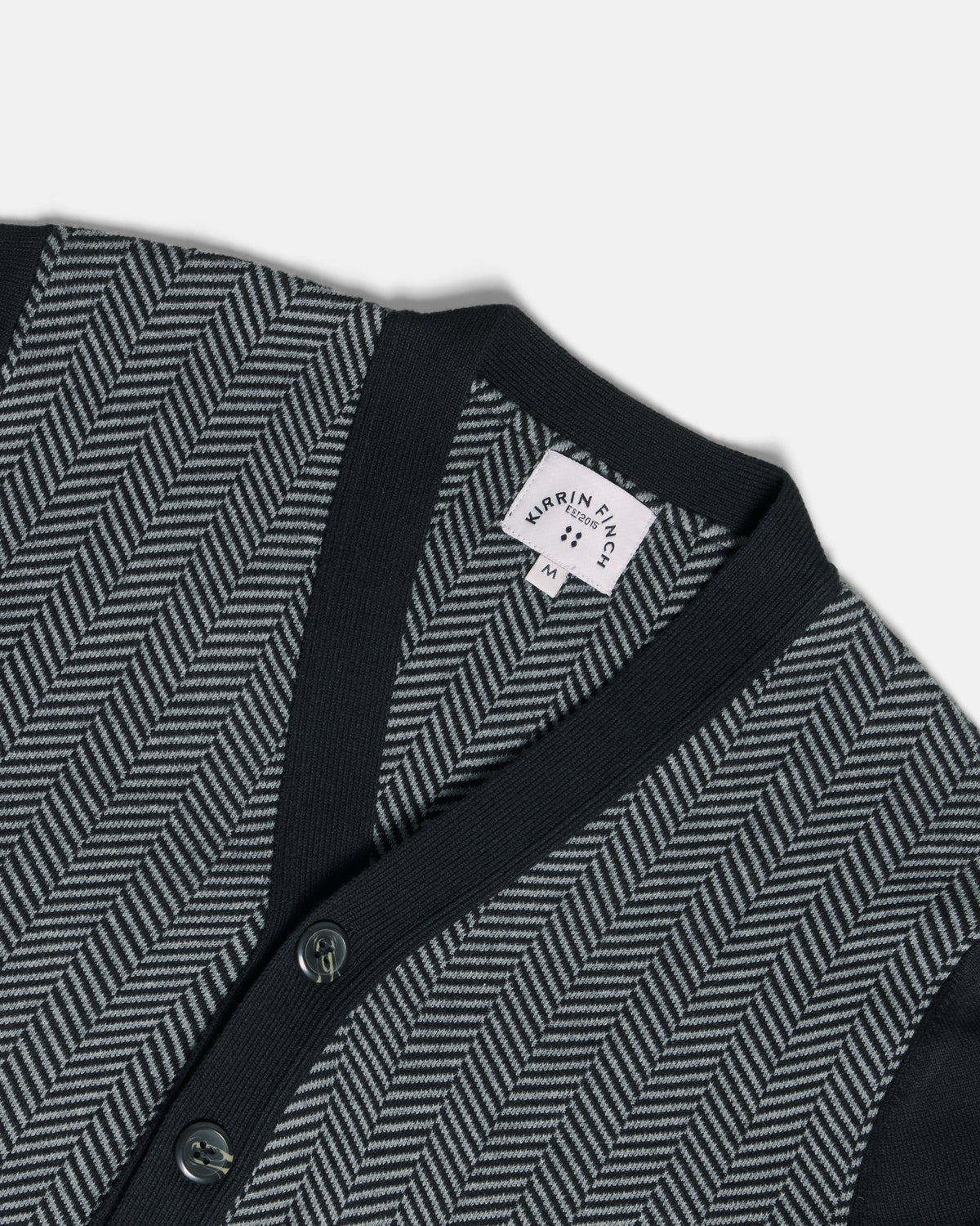 Close-up of a knit cardigan with a dark navy V-neck and button placket, paired with a gray and black herringbone pattern on the body. Color: Inkstone Herringbone