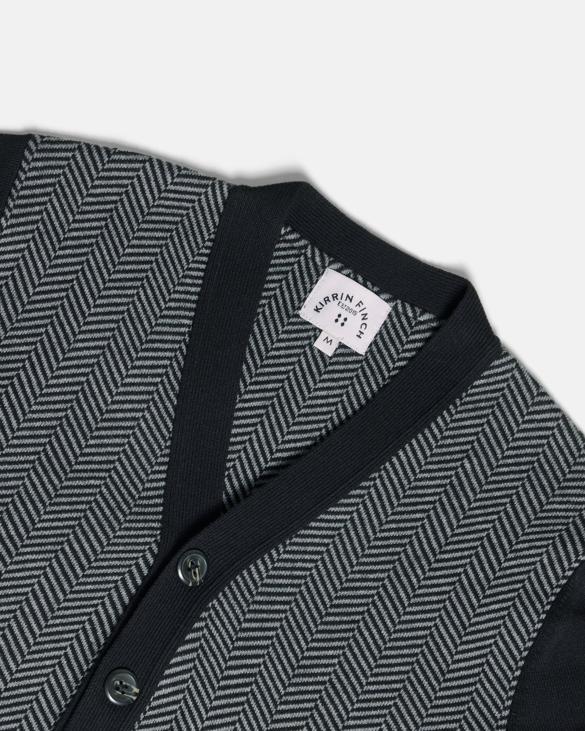 Close-up of a knit cardigan with a dark navy V-neck and button placket, paired with a gray and black herringbone pattern on the body. Color: Inkstone Herringbone