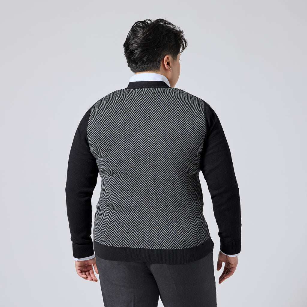 Model viewed from the back wearing a long-sleeve knit sweater with a black and white herringbone pattern on the body and solid black sleeves, collar, and hem, paired with dark trousers against a light gray studio background. Color: Inkstone Herringbone