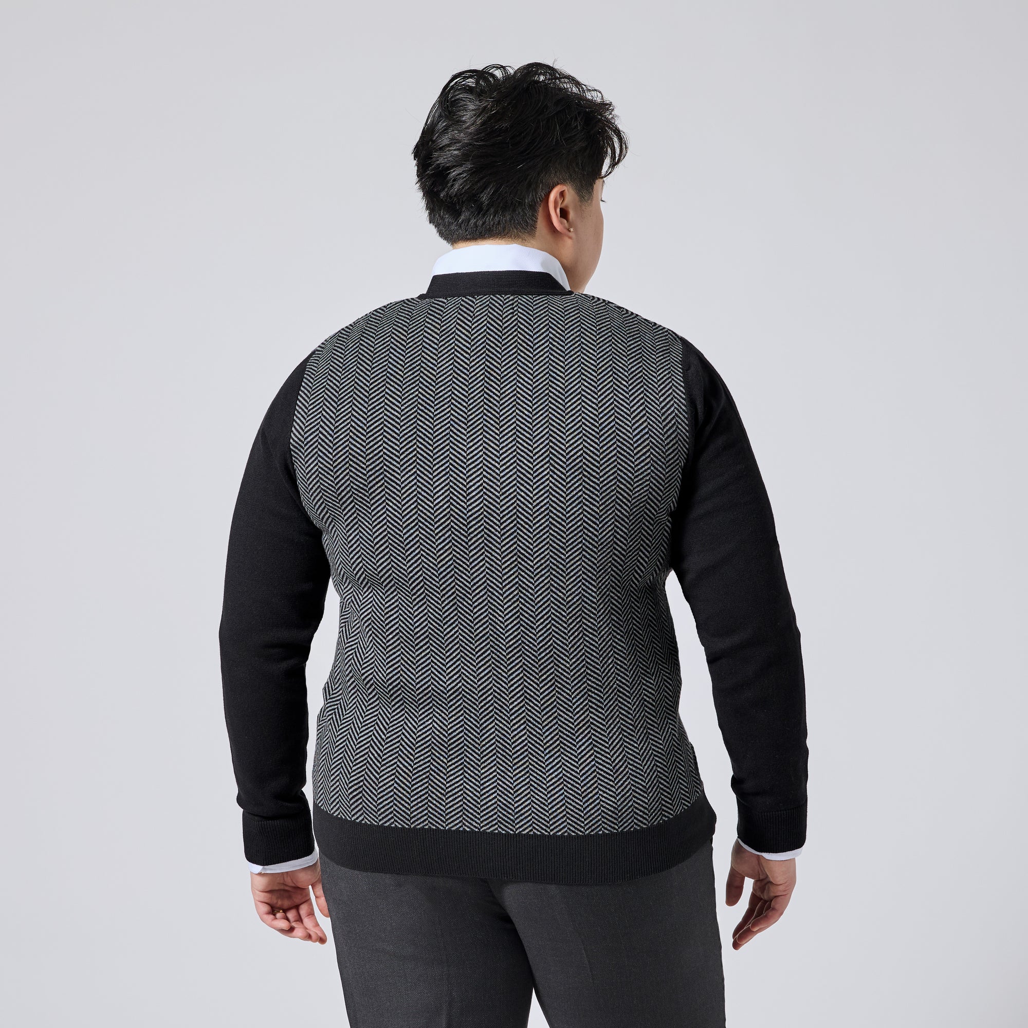 Model viewed from the back wearing a long-sleeve knit sweater with a black and white herringbone pattern on the body and solid black sleeves, collar, and hem, paired with dark trousers against a light gray studio background. Color: Inkstone Herringbone