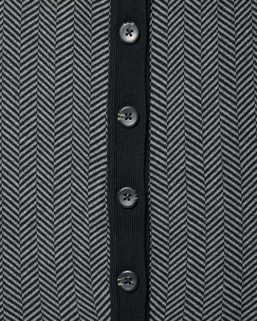 Close-up of a herringbone patterned knit garment in dark gray and black, featuring a solid black center placket with five dark buttons. Color: Inkstone Herringbone