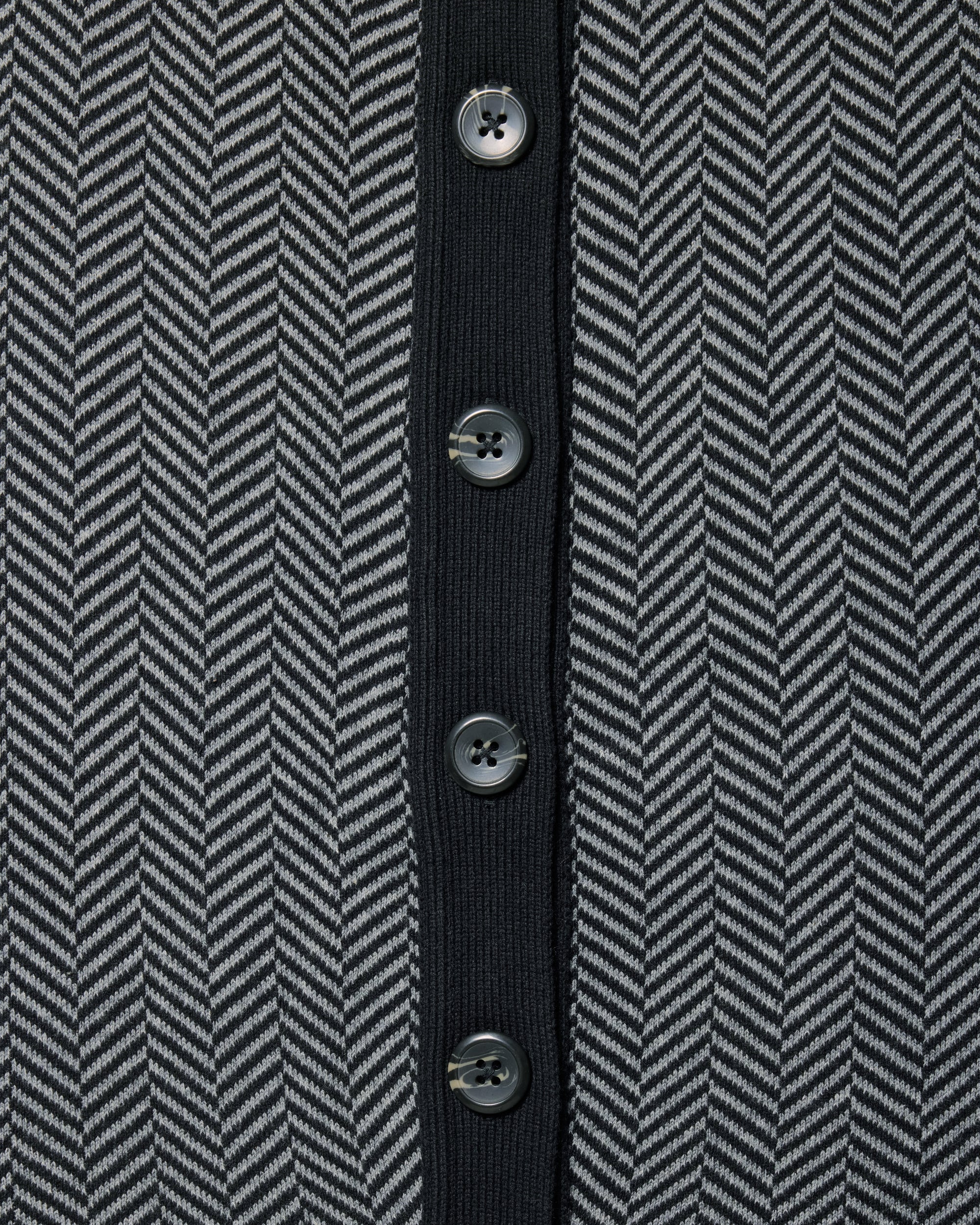Close-up of a herringbone patterned knit garment in dark gray and black, featuring a solid black center placket with five dark buttons. Color: Inkstone Herringbone