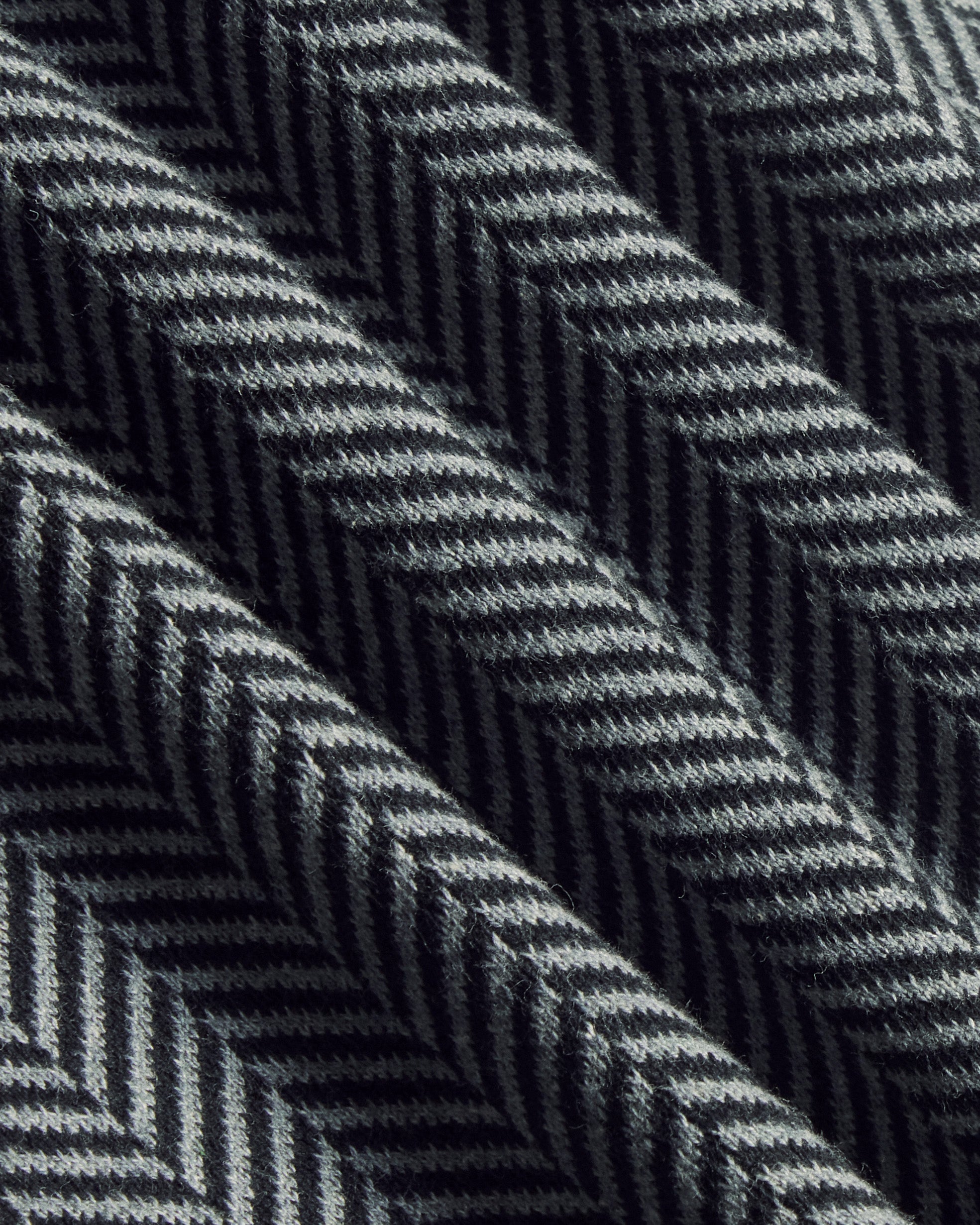 A close-up of a woven fabric with a dark gray and black herringbone pattern, showing the texture and diagonal folds. Color: Inkstone Herringbone