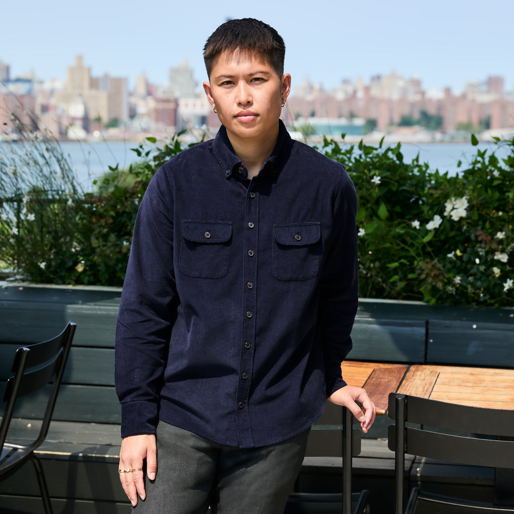 Model wearing a dark navy corduroy button-down shirt with two chest pockets, standing outdoors on a patio with a city skyline and water in the background. Color: Maritime Blue