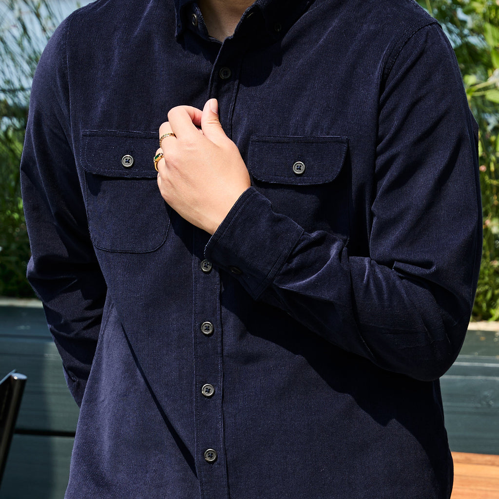 A close-up of a person wearing a dark navy corduroy long-sleeve button-up shirt with two chest pockets and metal buttons, set against an outdoor background with green foliage. Color: Maritime Blue