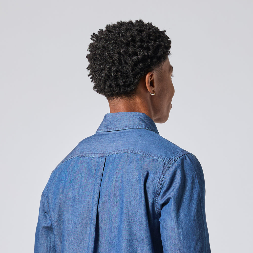 A person shown from the back wearing a blue denim button-up shirt against a light gray studio background. Color: Denim