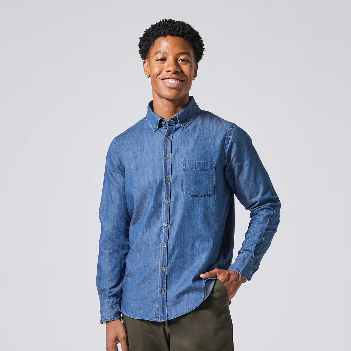 A smiling model wearing a medium blue long-sleeve button-down shirt with a chest pocket, paired with olive green pants, standing against a light gray studio background. Color: Denim