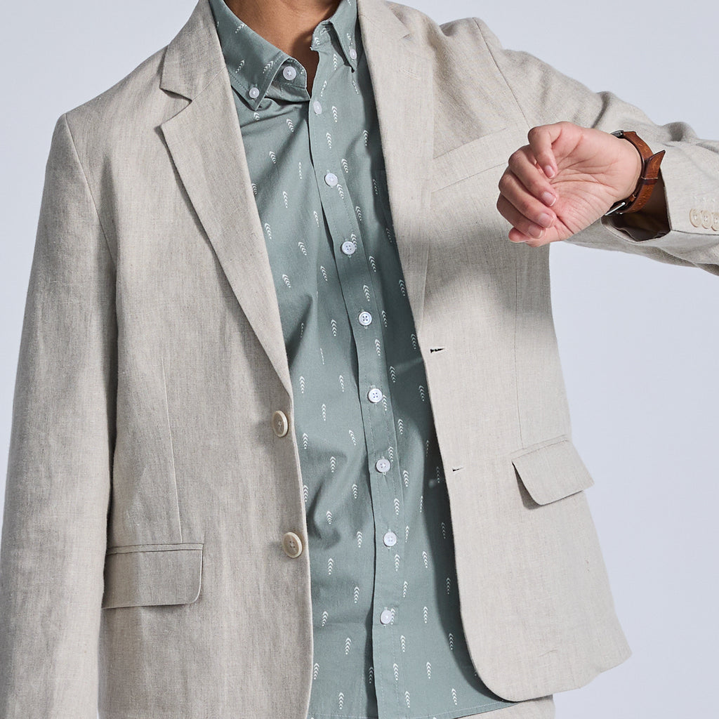 Model wearing linen blazer over a grween shirt looking at the watch on their wrist. Color: Oat