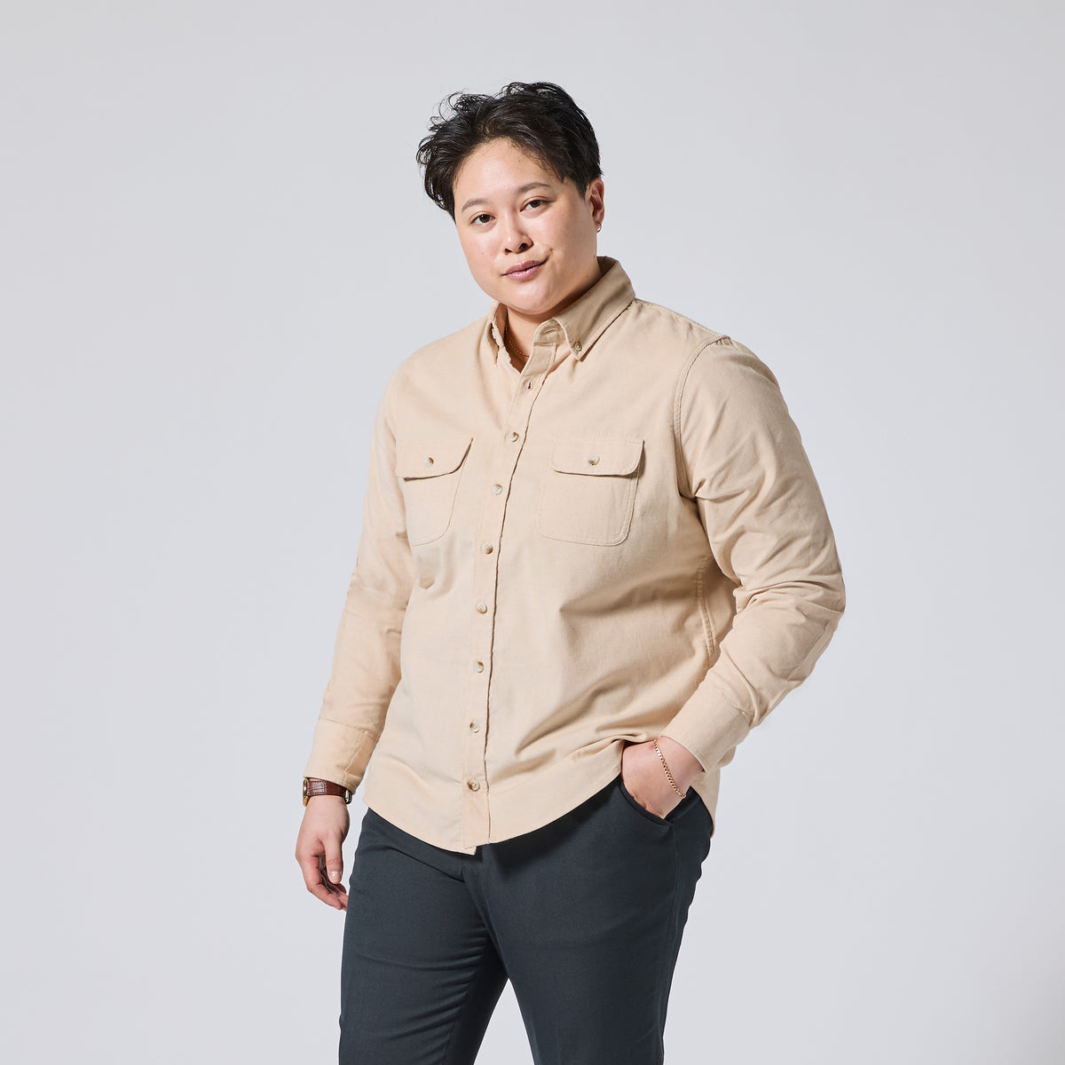 Model wearing a beige long-sleeve button-up shirt with two chest pockets and dark pants, standing against a light gray studio background. Color: Oatmeal
