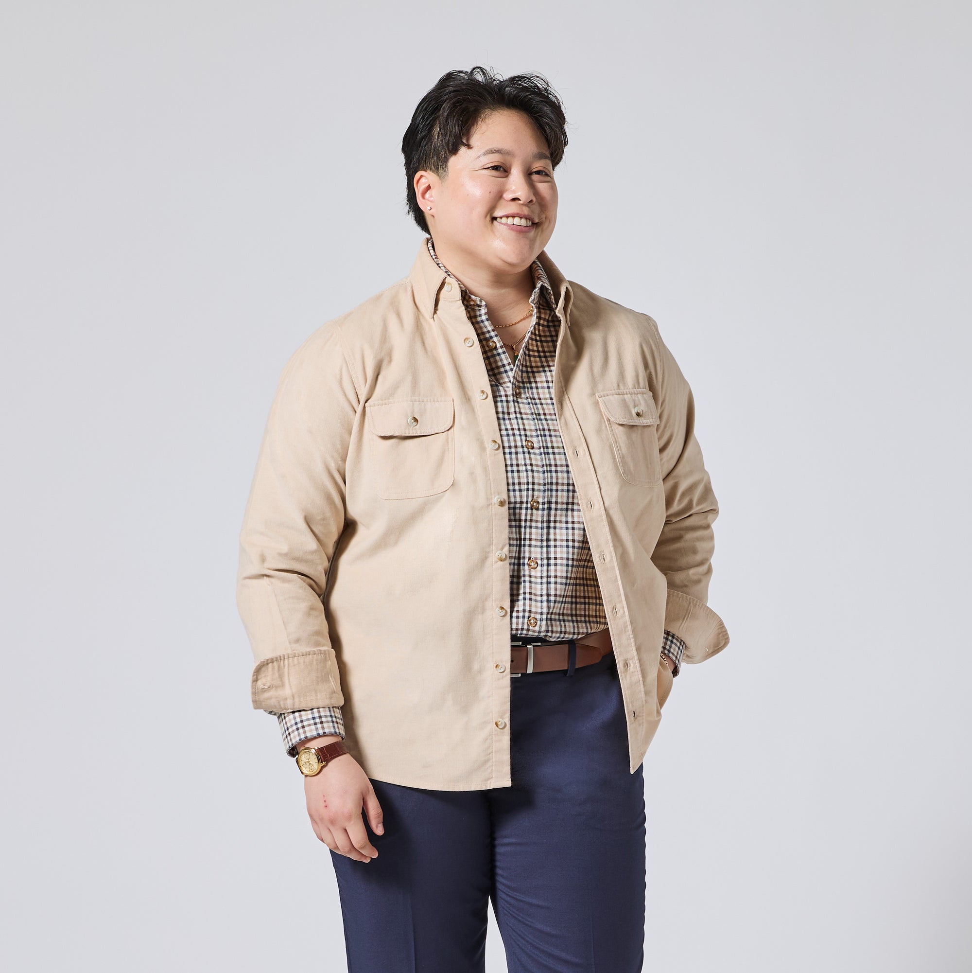 Model wearing a beige button-up overshirt over a brown and white plaid shirt, paired with navy blue trousers, standing against a light gray studio background. Color: Oatmeal