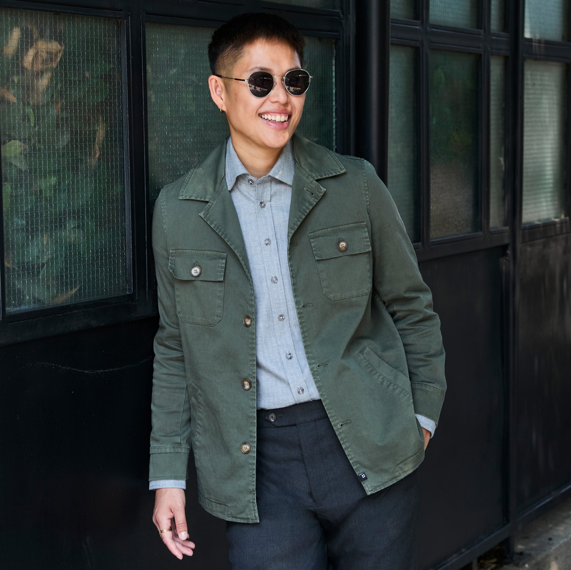 Model wearing a green utility jacket over a light blue button-down shirt and dark trousers, standing in front of a black metal building with windows. Color: Olive