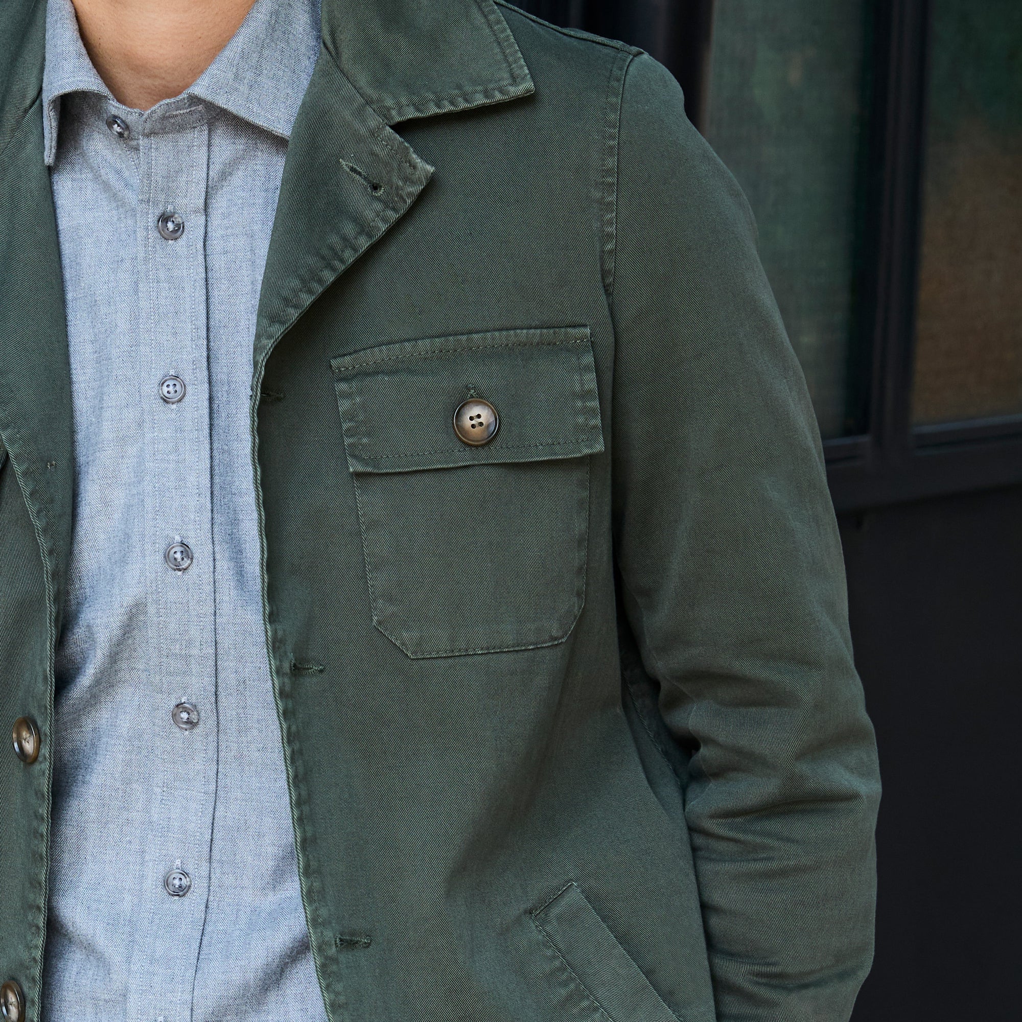 Close-up of a person wearing an olive green utility jacket over a light blue button-down shirt, with a dark window background. Color: Olive