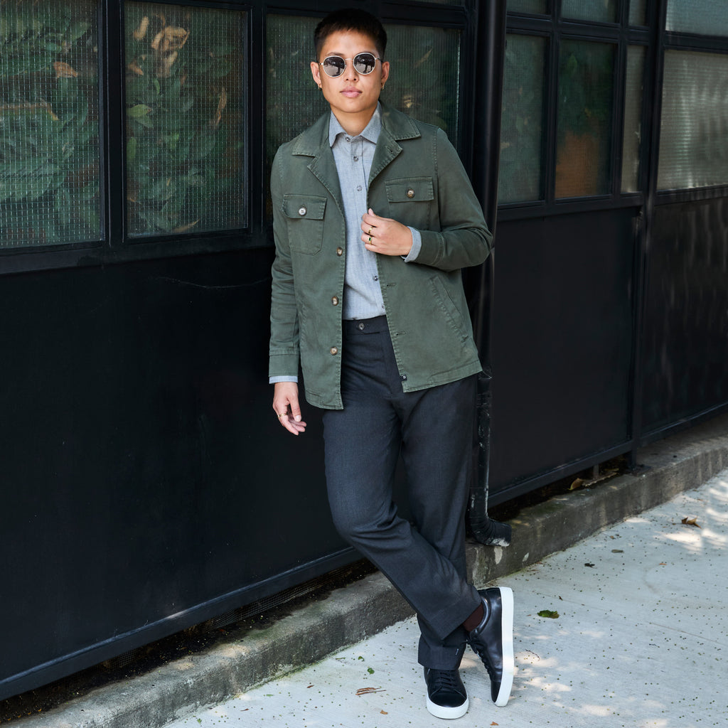 Model wearing an olive green utility jacket over a light gray button-down shirt, paired with dark charcoal pants and black sneakers, standing outdoors against a black metal building. Color: Olive
