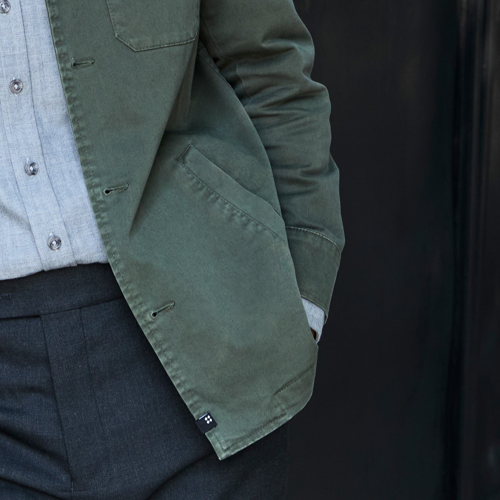 Close-up of a person wearing an olive green jacket over a light gray button-up shirt and dark charcoal trousers, with the jacket sleeve and chest pocket visible against a dark background. Color: Olive