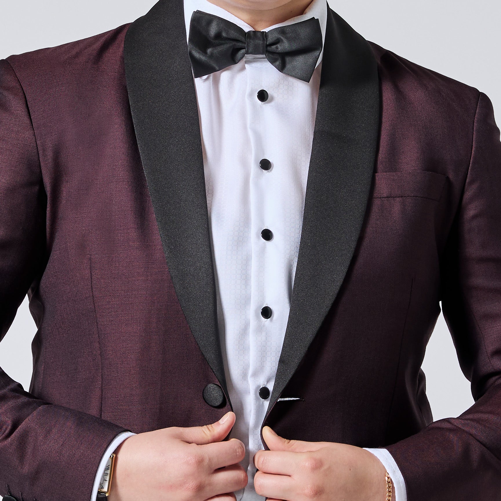 A close-up of a model wearing a formal tuxedo jacket in deep burgundy with black satin lapels, a white tuxedo shirt, black buttons, and a matching black bow tie. Color: Deep Oxblood