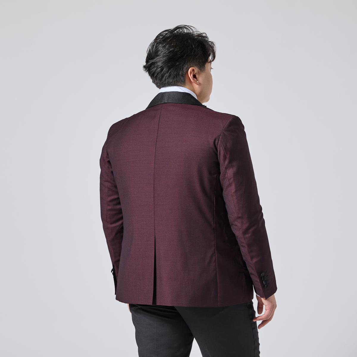 Model viewed from behind wearing a dark burgundy blazer with a black collar, paired with a white shirt and dark trousers, against a light gray studio background. Color: Deep Oxblood