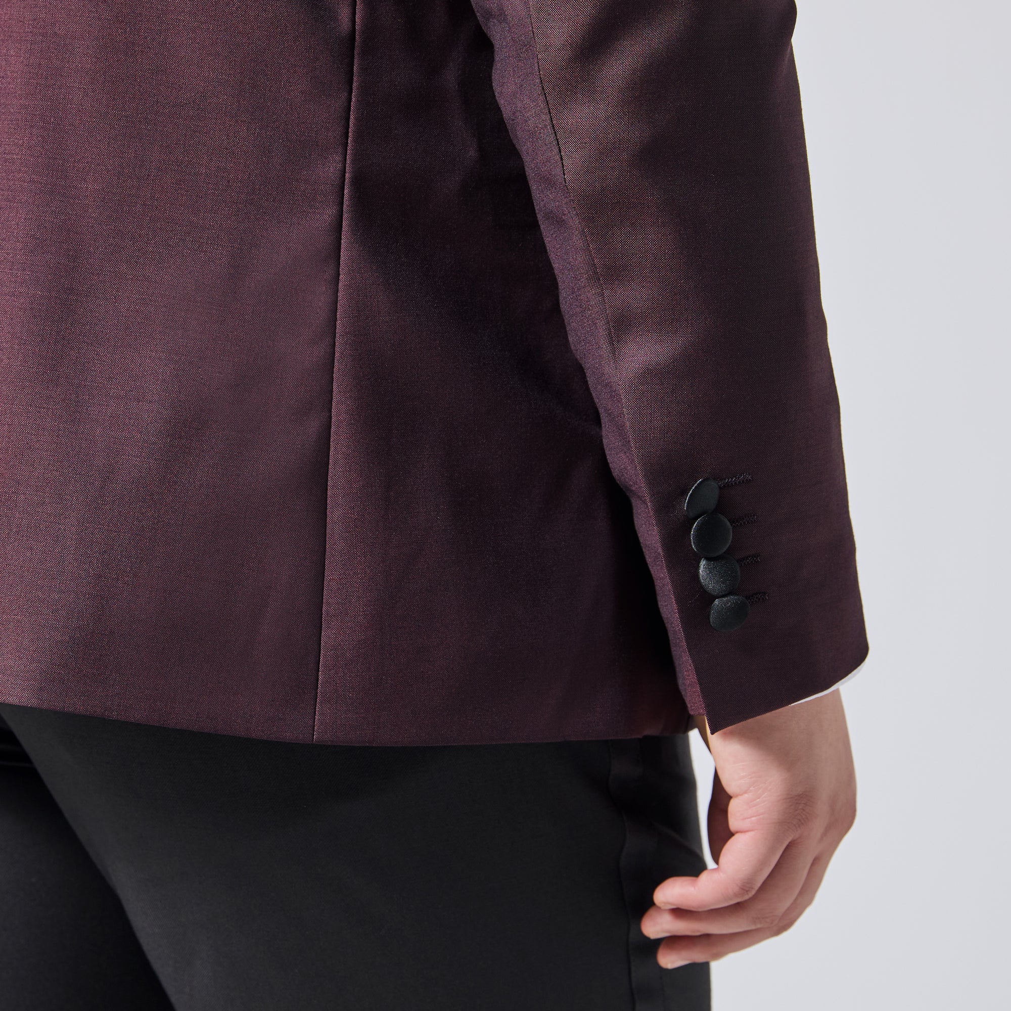 Close-up of a person wearing a dark plum blazer with three black buttons on the cuff, paired with black trousers, against a light gray background. Color: Deep Oxblood