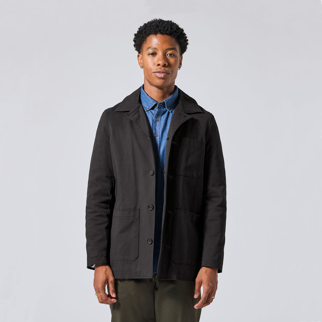 Model wearing a black utility jacket over a blue button-down shirt and olive green pants, standing against a light gray studio background.