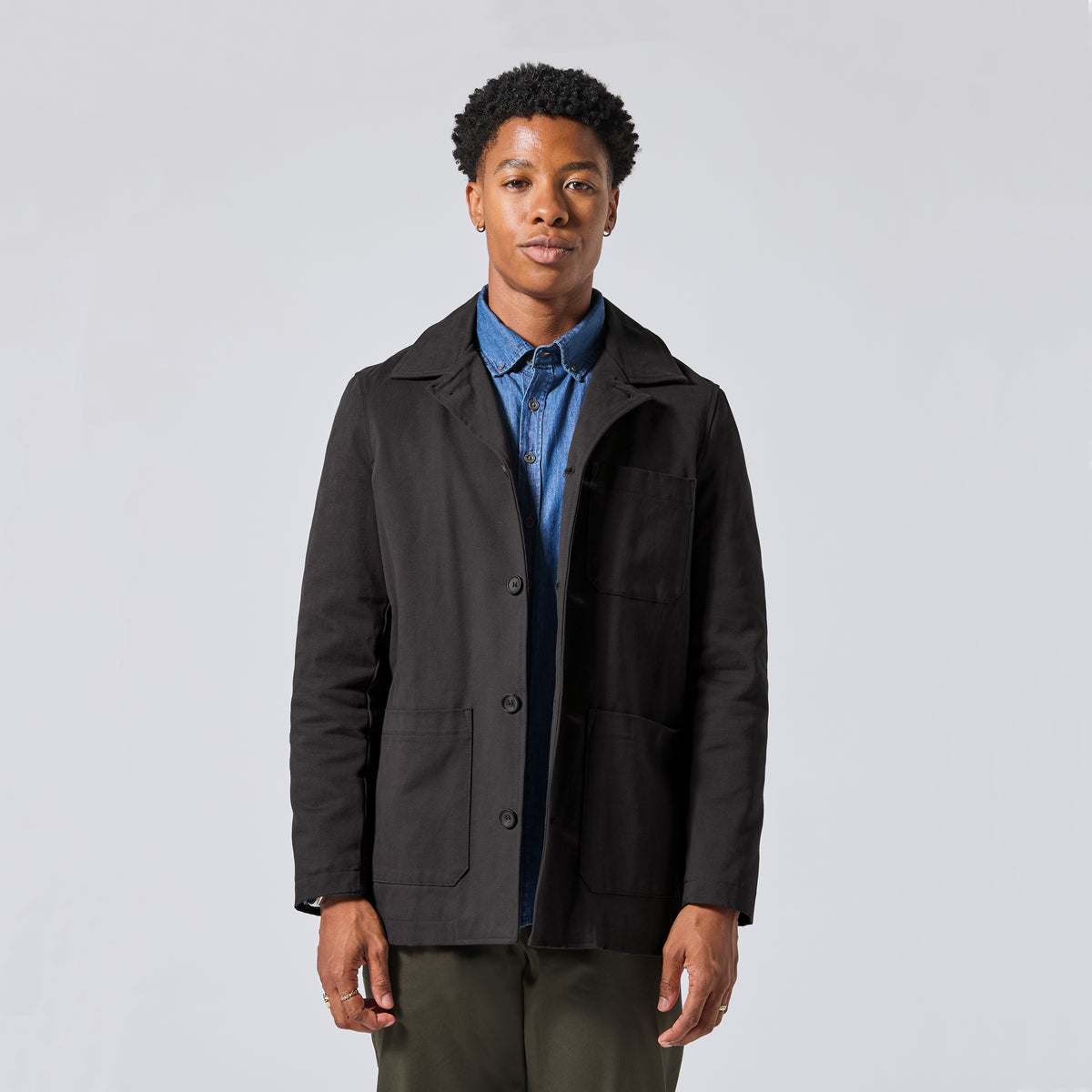 Model wearing a black utility jacket over a blue button-down shirt and olive green pants, standing against a light gray studio background.