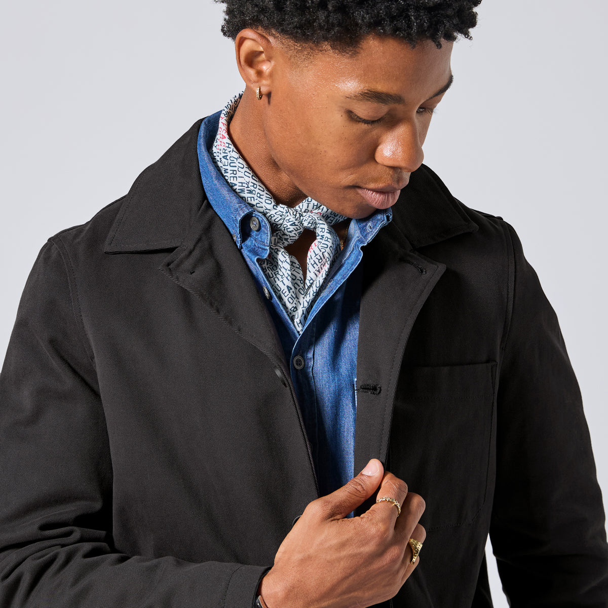 Model wearing the Raven Quilted Jacket over a blue button-up shirt and a patterned neck scarf, shown from the chest up against a light gray studio background.