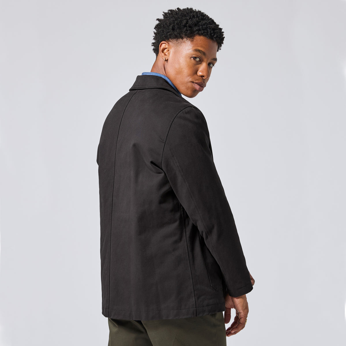 Model wearing the Raven Quilted Jacket over a blue collared shirt and olive green trousers, photographed from the back-three-quarter angle against a light gray studio background.