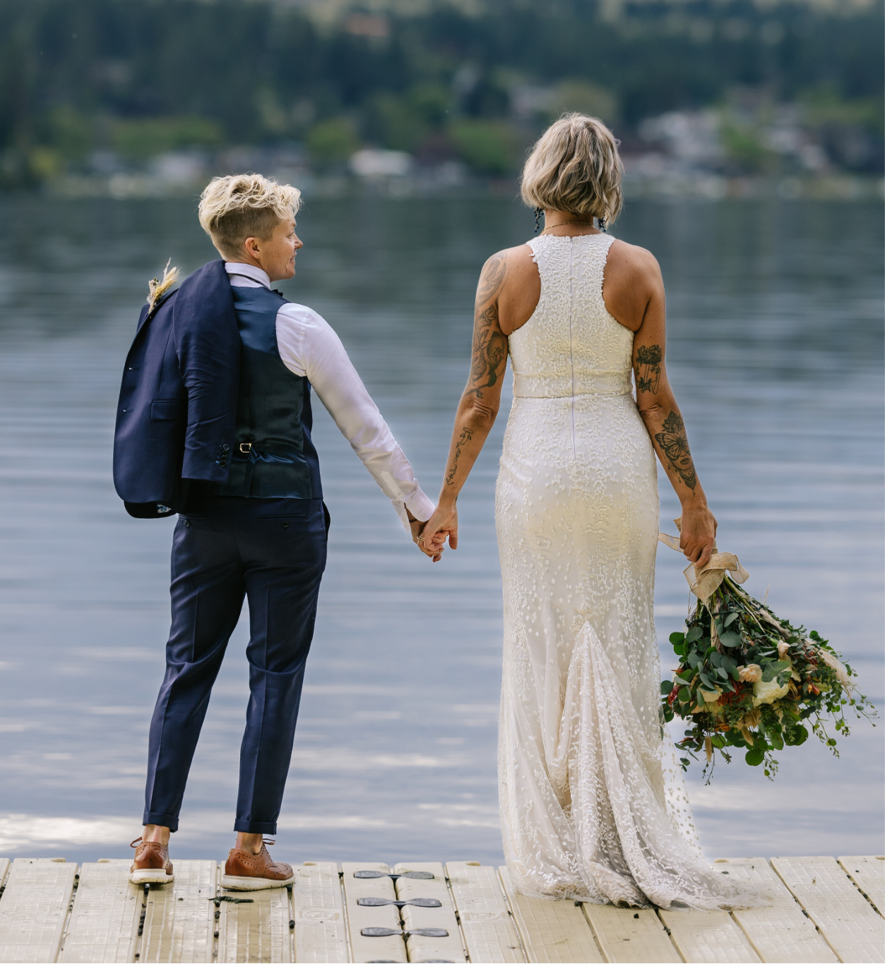 Two people ( Two brides) holding hands on a dock by a lake. The person on the left is wearing a vest, dress pants, dress shirt and has her blazer tossed over her shoulder. Person on right is wearing a white wedding dress with flowers in her other hand. 