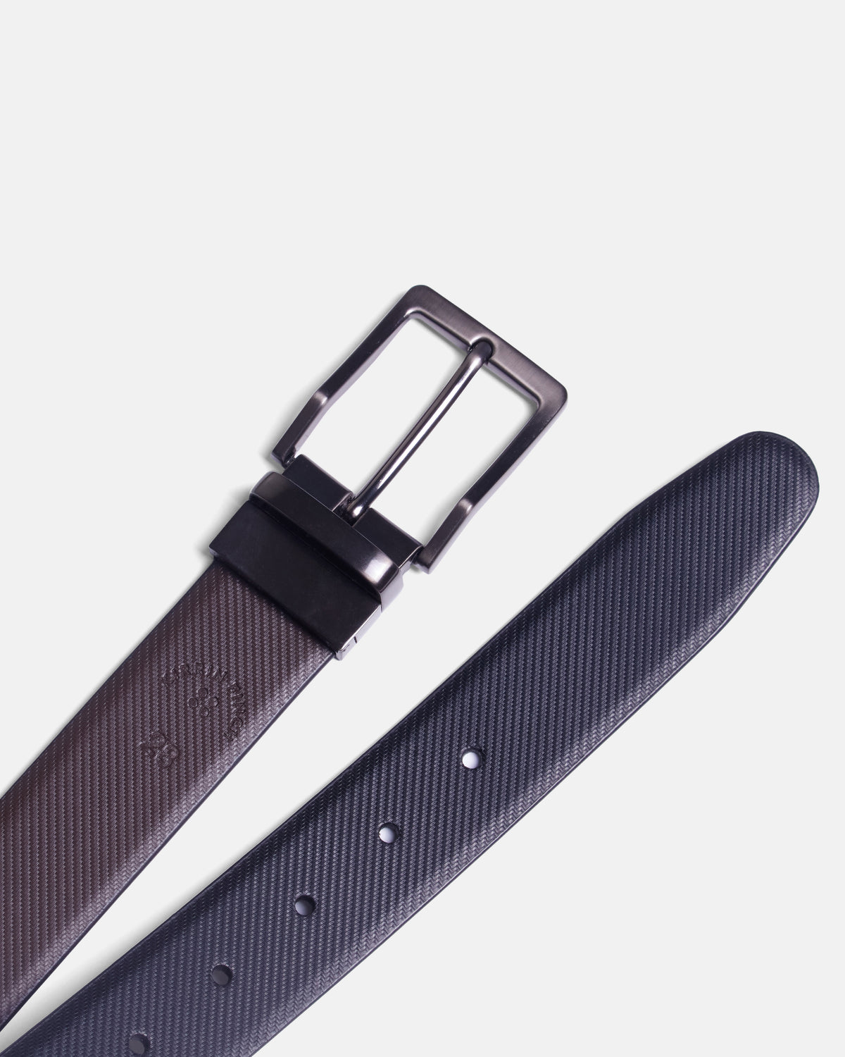 A close-up of a textured leather belt with a dark metallic buckle, shown on a light gray background. Color: Black Twill/Brown Twill