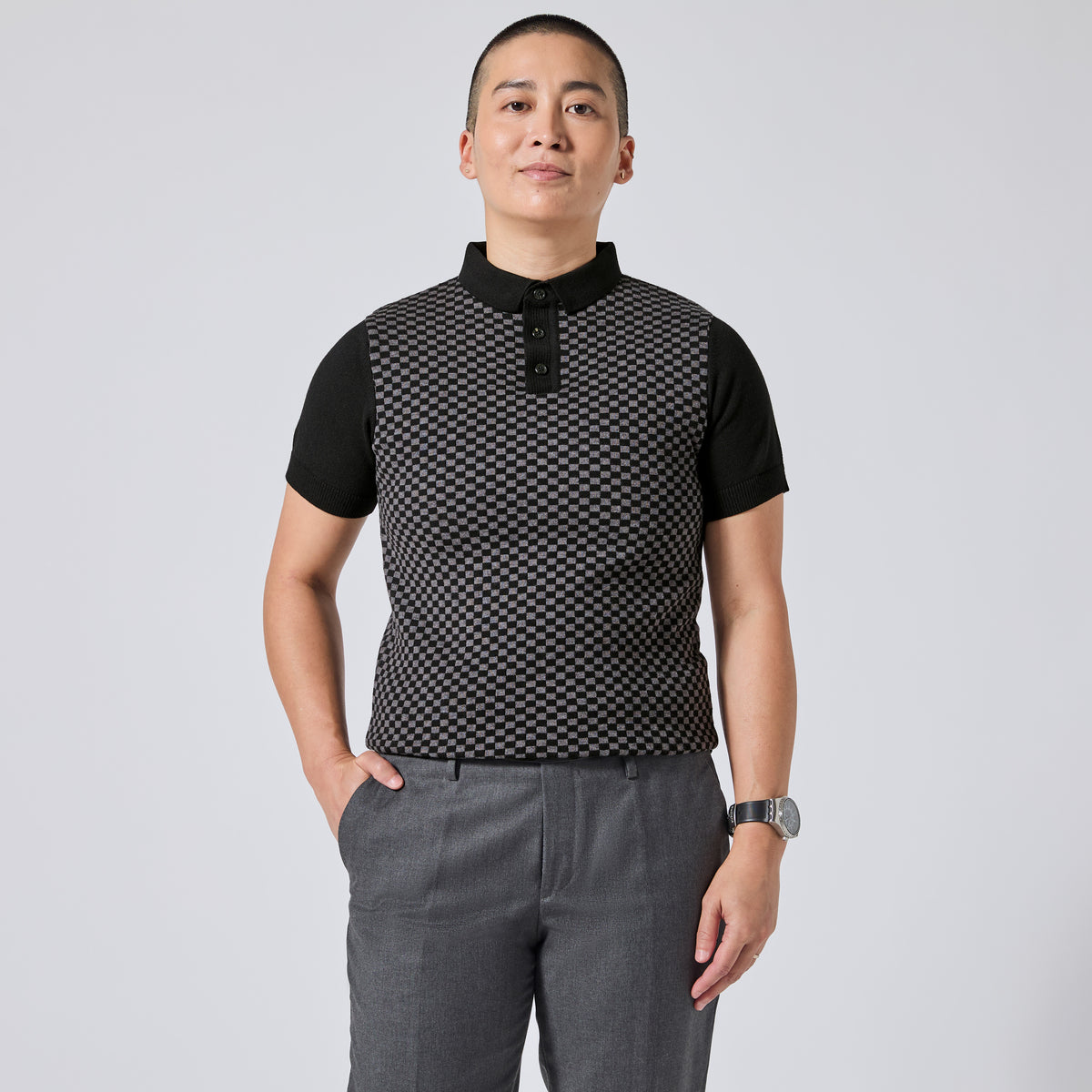 Model wearing a black short-sleeve polo shirt with a textured black-and-white geometric pattern on the front panel, paired with gray trousers, standing against a light gray studio background. Color: Shadow Checker
