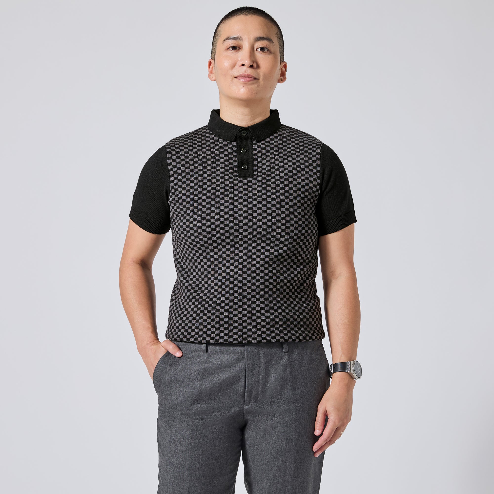Model wearing a black short-sleeve polo shirt with a textured black-and-white geometric pattern on the front panel, paired with gray trousers, standing against a light gray studio background. Color: Shadow Checker
