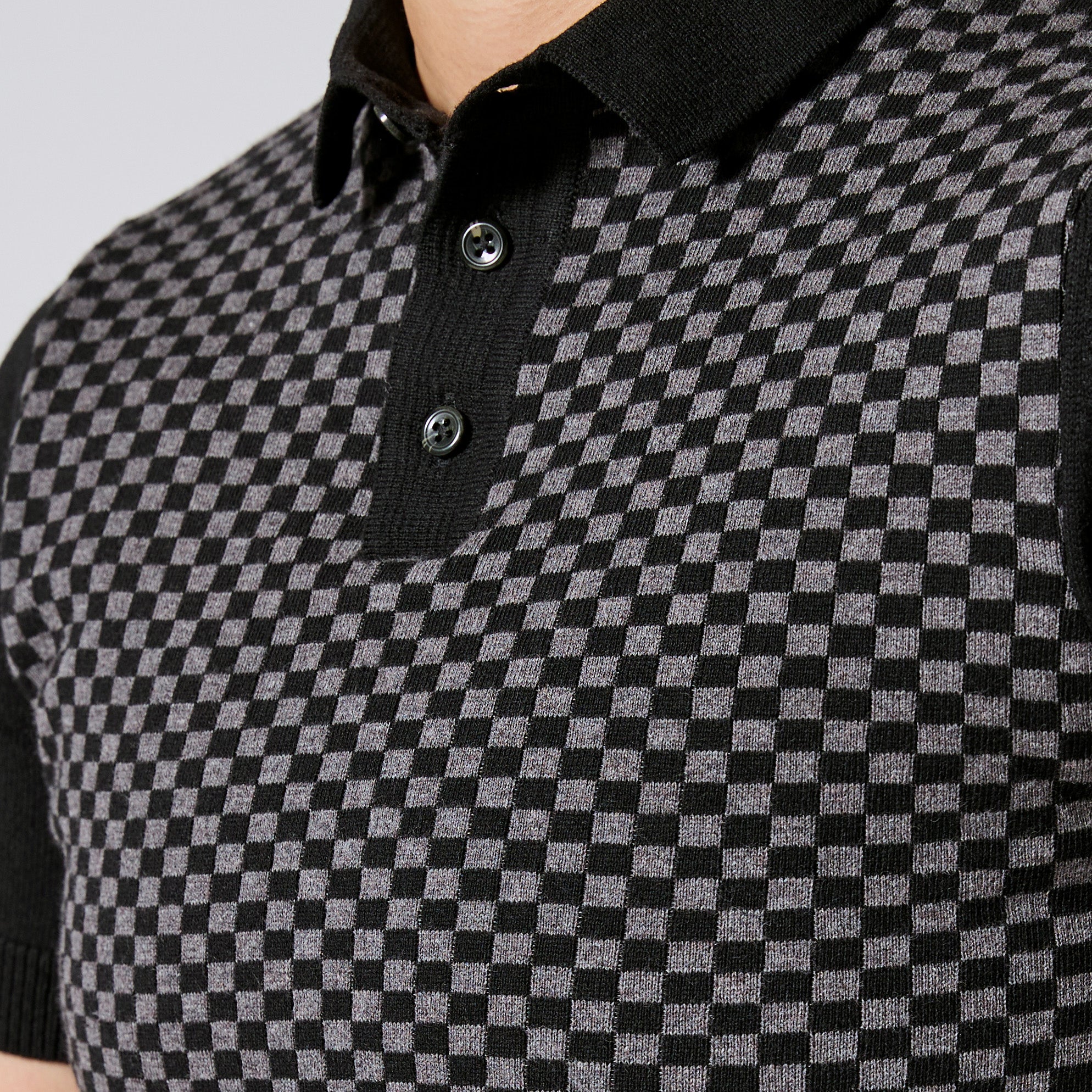 Close-up of a black and gray checkered knit polo shirt with a black collar and two dark buttons. Color: Shadow Checker