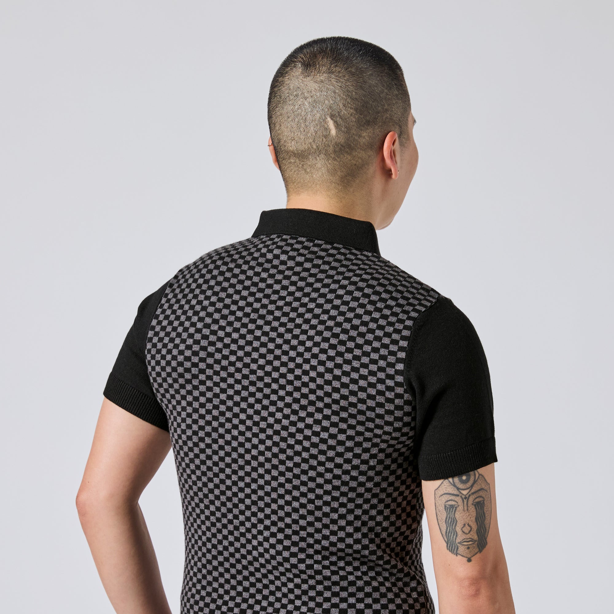Model viewed from behind wearing a short-sleeve polo shirt with a black and gray checkerboard pattern on the back and solid black sleeves, collar, and cuffs.  Color: Shadow Checker