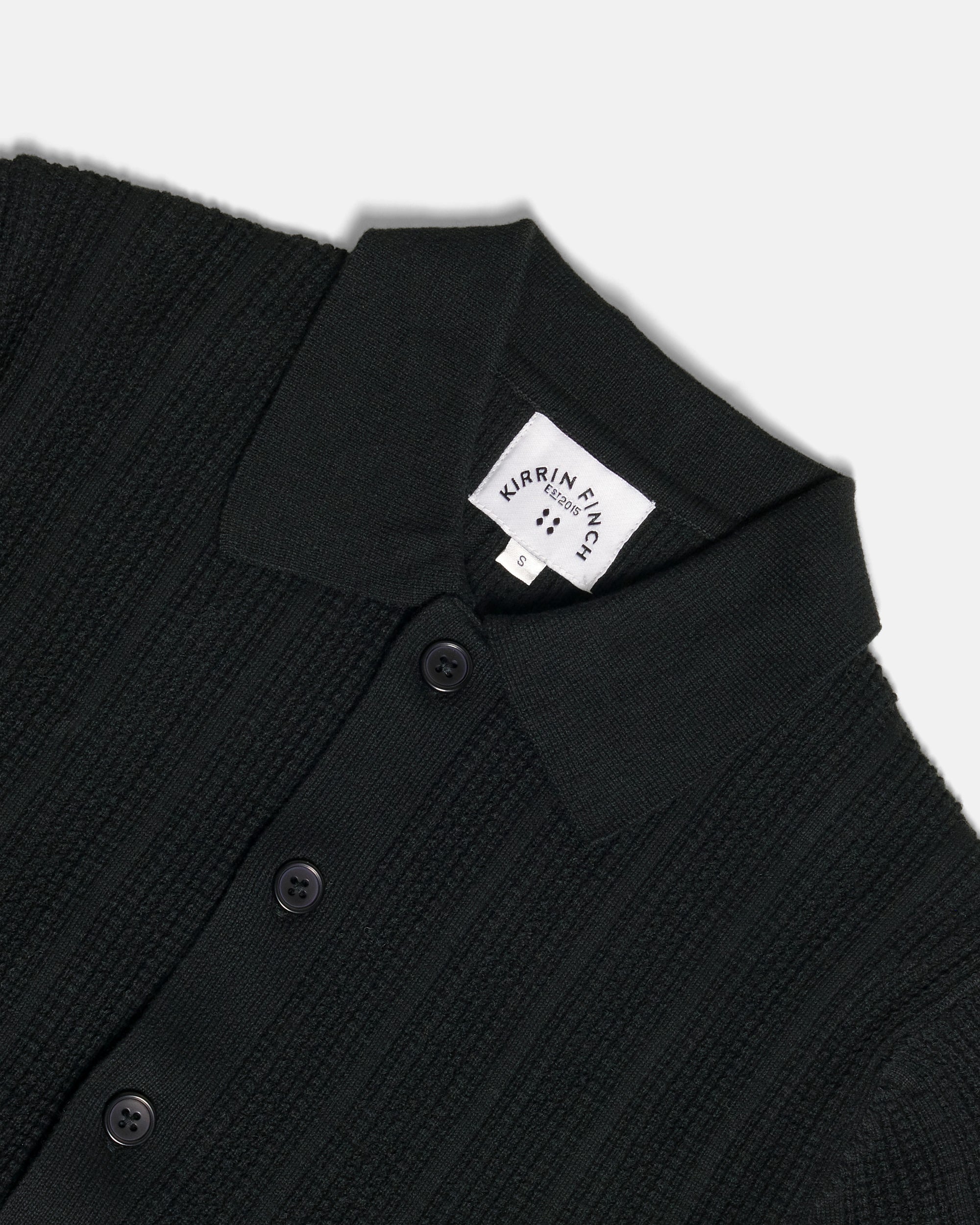 Close-up of a black ribbed knit polo shirt by Kirrin Finch, showing the collar with a white brand label and dark buttons on a light gray background. Color: Black