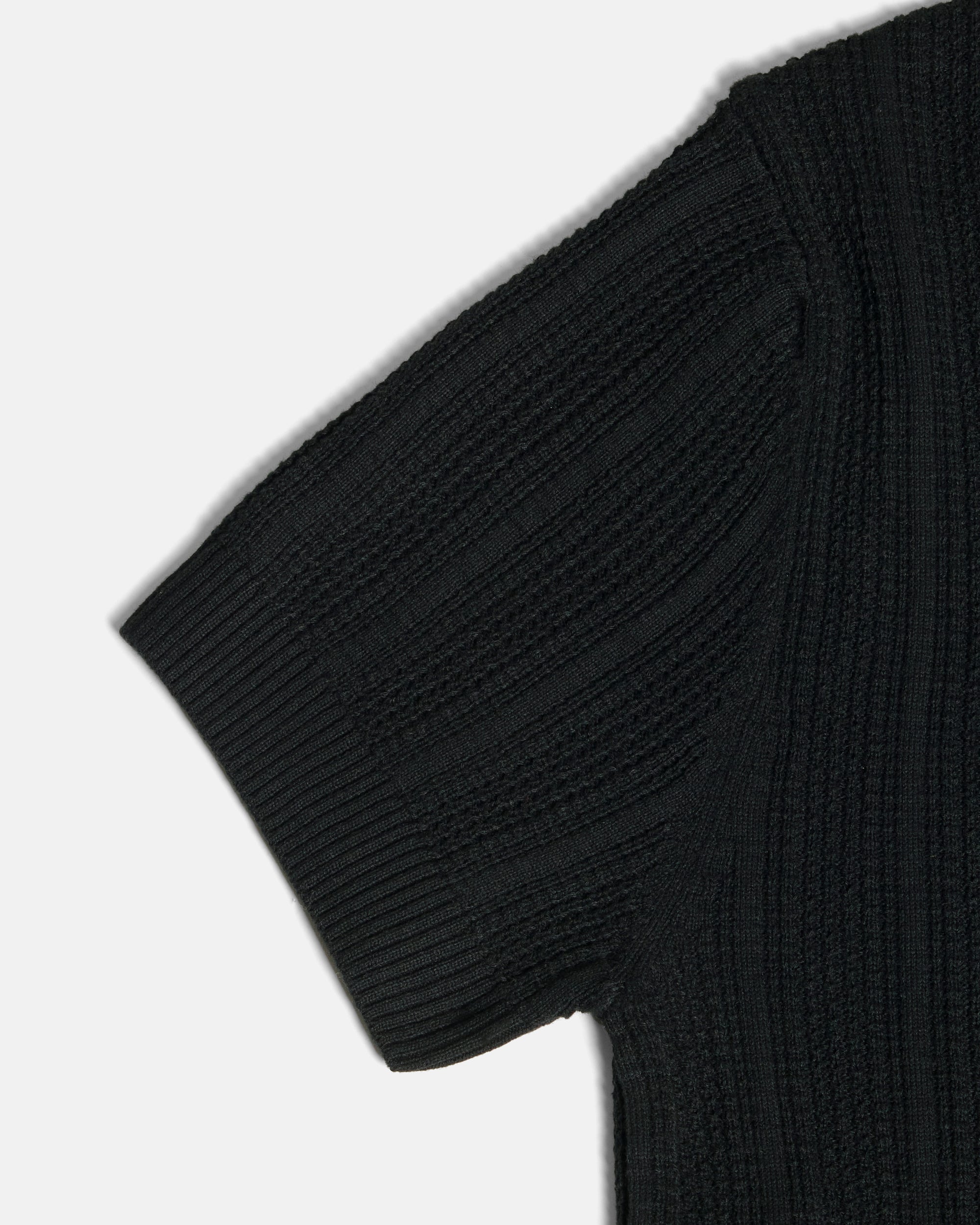 Close-up of a black ribbed knit short-sleeve top showing the sleeve and shoulder area against a light gray background. Color: Black