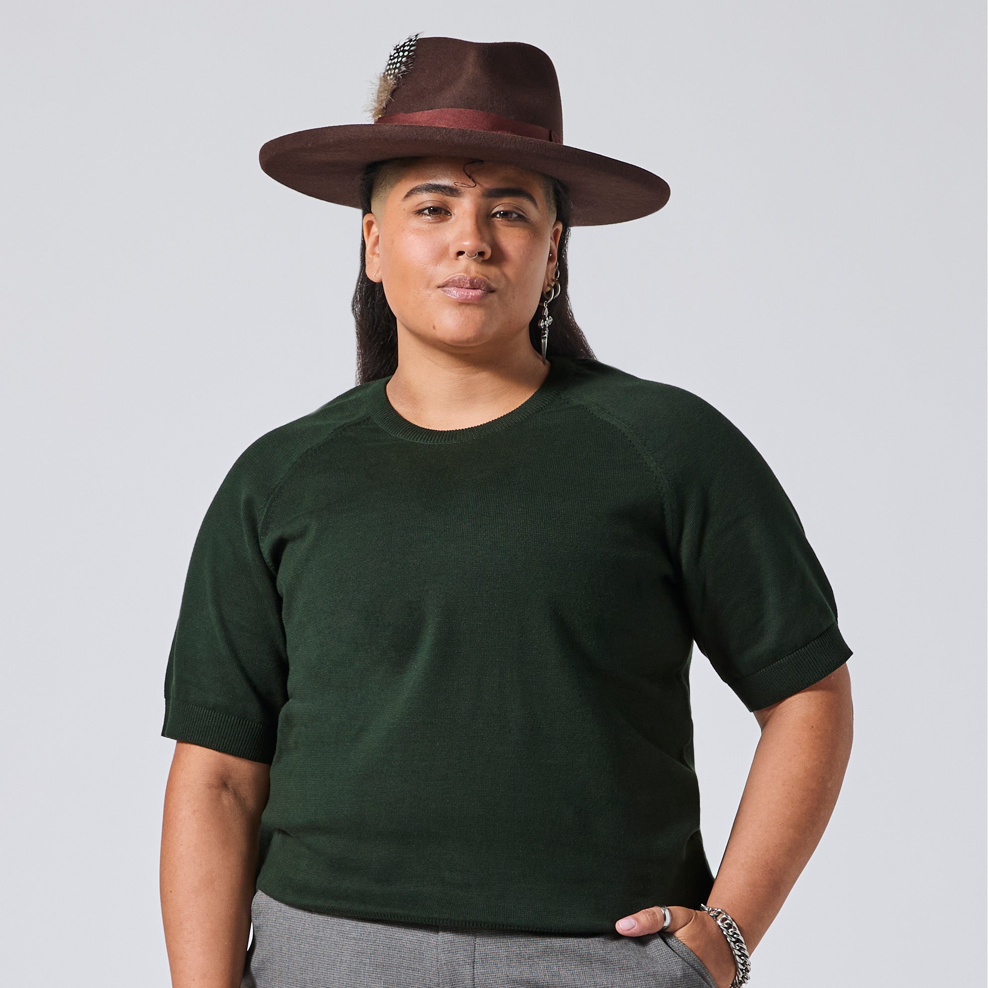 A person wearing a dark forest green short-sleeve crewneck t-shirt, a brown wide-brim hat with a feather, and gray pants, standing against a light gray studio background. Color: Evergreen