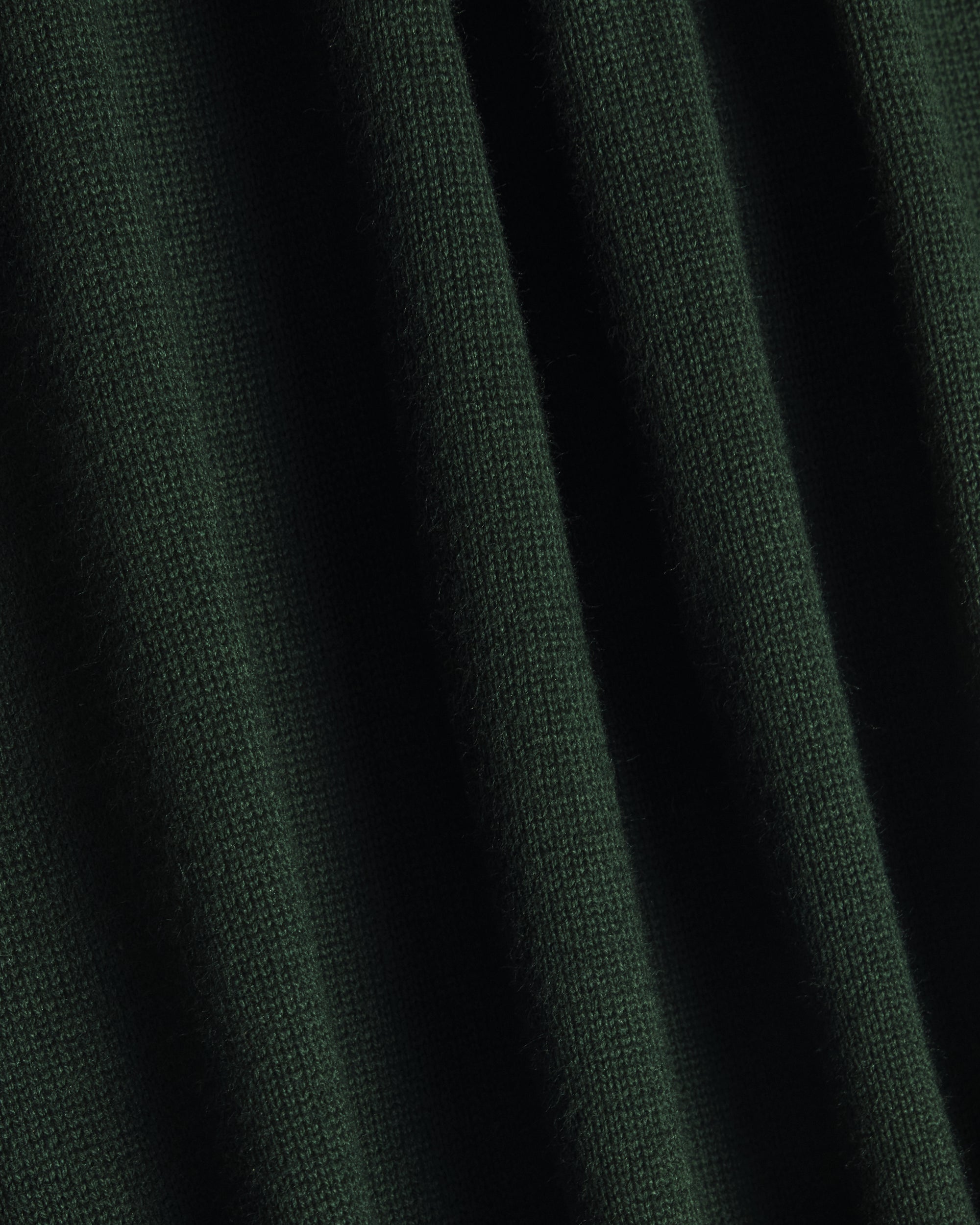 A close-up of dark forest green knit fabric with soft vertical folds showing the texture and weave. Color: Evergreen