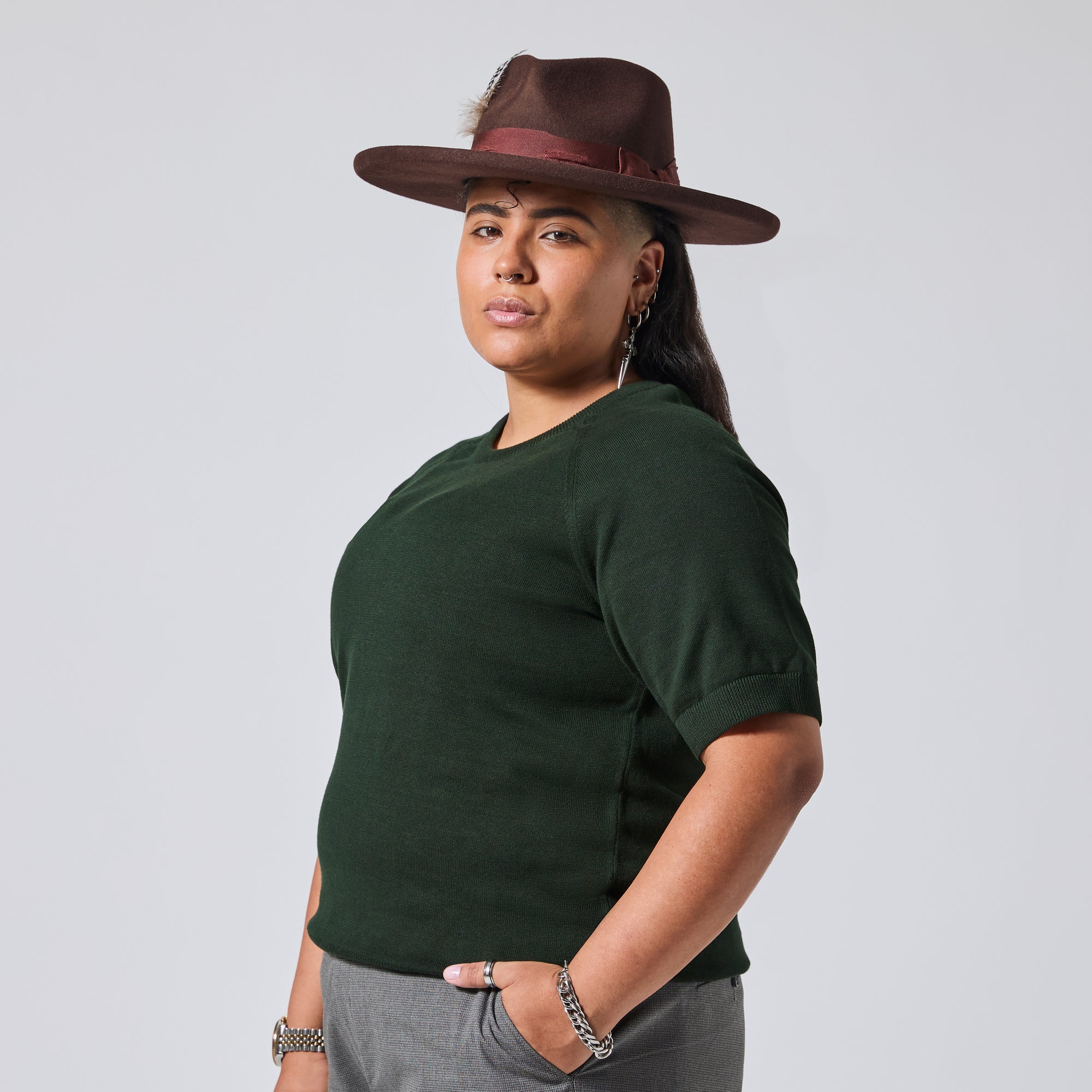 A model wearing a dark forest green short-sleeve knit top, gray pants, and a brown wide-brim hat, photographed against a light gray studio background. Color: Evergreen