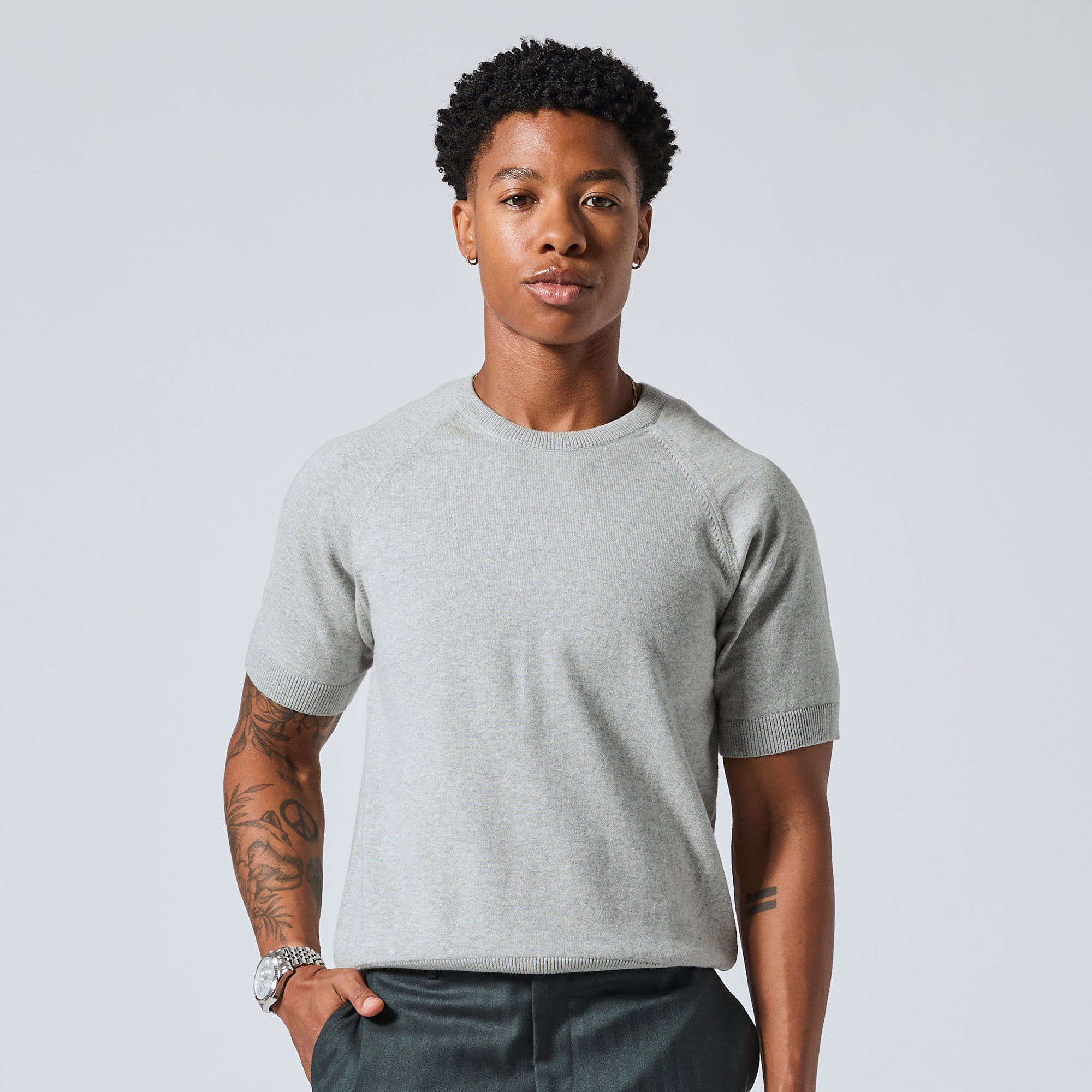 Model wearing a light gray short-sleeve crewneck t-shirt and dark gray pants, standing against a light gray studio background. Color: Heather Grey