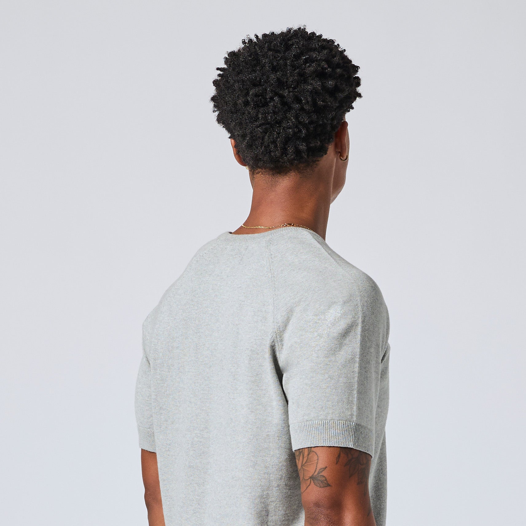 Model viewed from behind wearing a light gray short-sleeve knit t-shirt, showcasing the back collar and shoulder construction against a light gray background. Color: Heather Grey
